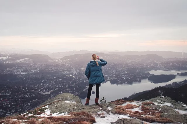 A woman stands on a mountain peak, gazing at a serene lake and surrounding mountains.