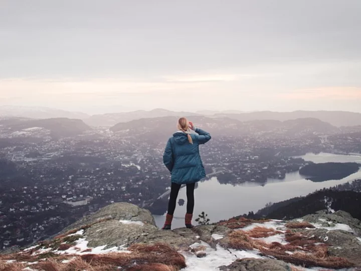 A woman stands on a mountain peak, gazing at a serene lake and surrounding mountains.